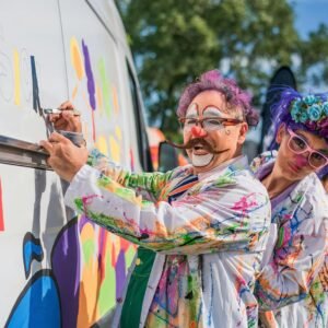 Clowns in colorful costumes painting a van at a fun outdoor event under sunny skies.