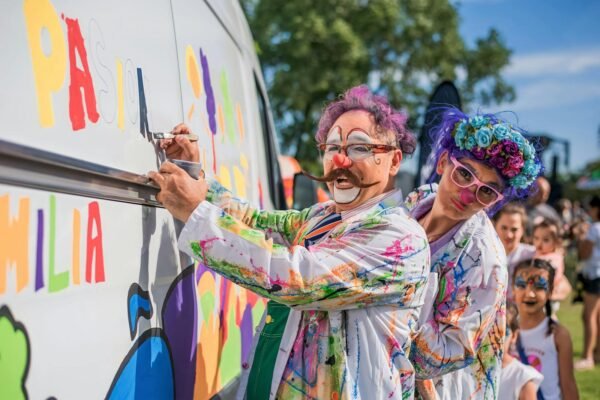 Clowns in colorful costumes painting a van at a fun outdoor event under sunny skies.